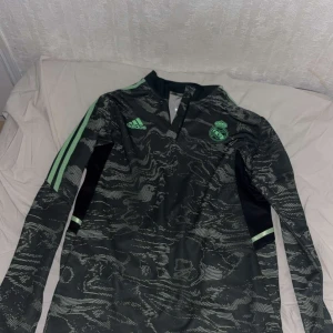 Svart och grön fotbollströja från Adidas - Säljer en snygg svart och grön långärmad fotbollströja från Adidas med ett unikt mönster. Tröjan har tre gröna ränder längs ärmarna och Real Madrid på bröstet. Perfekt för fotbollsfans som vill ha något stilrent och sportigt.