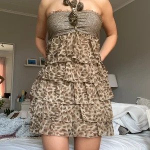Brun axelbandslös klänning med volanger - Säljer en snygg brun axelbandslös klänning med volanger och leopardmönster. Överdelen är glittrig med en dekorativ blomma och knytband. Perfekt för en festlig kväll!