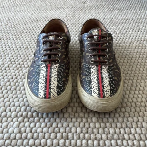 Burberry sneakers - Snygga bruna sneakers från Burberry med ett iögonfallande monogrammönster. Skorna har en röd och vit rand längs mitten och klassisk snörning. Perfekta för att ge din outfit en lyxig touch. Enda skadan är en spricka vid fotöppningen som enkelt kan fixas hos en skomakare eller limma ihop själv. Nypris 4300