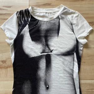 Vit och svart t-shirt med tryck - Säljer en cool vit t-shirt med ett svart tryck som täcker framsidan. T-shirten har korta ärmar och en rund halsringning. Perfekt för en unik och stilren look.