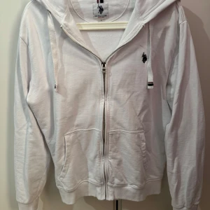 Vit hoodie från U.S. Polo Assn. - Säljer en stilren vit hoodie från U.S. Polo Assn. med dragkedja och broderad logga på bröstet. Den har en praktisk huva med snören och två fickor framtill. Perfekt för en avslappnad look.