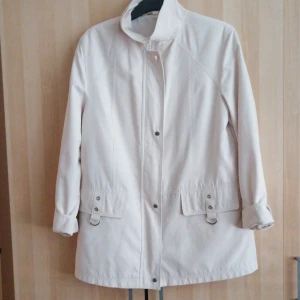 Beige kappa med knappar - Säljer en stilren beige kappa med knappar framtill och justerbara ärmslut. Jackan har praktiska fickor med dekorativa spännen och en hög krage för extra stil. Perfekt för vår och höst!