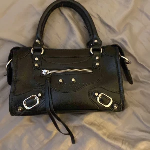 Svart handväska - Snygg svart handväska som är köpt på en marknad i Spanien. Använd ett fåtal gånger. Ett band som man kan sätta på så man kan ha den på axeln medkommer!