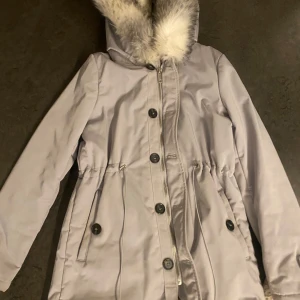 Ljusgrå jacka - Priset kan diskuteras! Snygg ljusgrå parkjacka med päls på luvan.  Den har även borttagbar päls vid dragkedjan. Jackan har både dragkedja och knappar framtill samt justerbar midja för en perfekt passform. Perfekt för kyliga dagar! Köptes för 4000. Den har tyvärr hål i fickan.