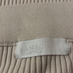 Beige ribbad kjol från Samsa Collection - Snygg beige ribbad kjol från Samsa Collection i storlek M. Kjolen har en stilren design med en slits baktill som ger extra rörelsefrihet. Perfekt för en chic och avslappnad look.