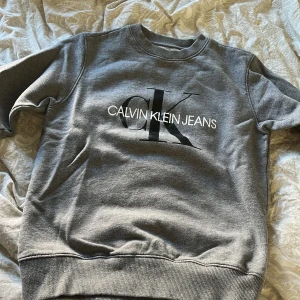 Grå sweatshirt från Calvin Klein Jeans - Säljer en stilren grå sweatshirt från Calvin Klein Jeans med deras ikoniska logga i svart och vitt på bröstet. Tröjan har en rund halsringning och ribbade muddar vid ärmslut och nederkant. Det är en herr s men passar som en dam m💕 säljes för den inte kommer till användning och är därmed i nyskick💕