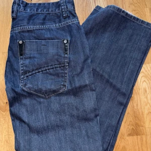 Blå jeansbyxor från Next - Snygga blå jeansbyxor från Next med klassisk femficksdesign och knappgylf. Perfekta för en avslappnad stil. De har en rak passform och är tillverkade i slitstarkt denim.