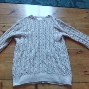Beige stickad tröja - Säljer en mysig beige stickad tröja med kabelstickat mönster. Tröjan har långa ärmar och ribbstickade muddar vid ärmslut och nederkant. Perfekt för kyliga dagar!