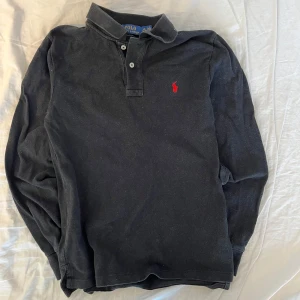 Svart pikétröja från Ralph Lauren - Säljer en stilren svart pikétröja från Ralph Lauren i storlek L Barnstorlek . Tröjan är tillverkad i 100% bomull och har en klassisk krage med knappar. Den har en röd broderad logotyp på bröstet och långa ärmar. Perfekt för en avslappnad men snygg look.