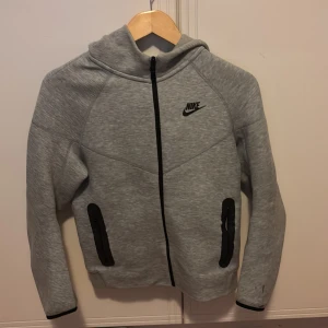 Grå träningsset från Nike - Säljer ett grått träningsset från Nike med en långärmad hoodie och matchande byxor. Hoodien har dragkedja och Nike-logga på bröstet. Byxorna har snörning i midjan och fickor med dragkedja. Perfekt för träning eller en avslappnad dag.