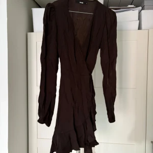 Brun omlottklänning från Bik Bok - Snygg brun skinrig omlottklänning från Bik Bok i storlek S. Klänningen har långa ärmar och volangdetaljer vid fållen. Perfekt för en stilren look. Mycket tunn och perfekt till sommaren