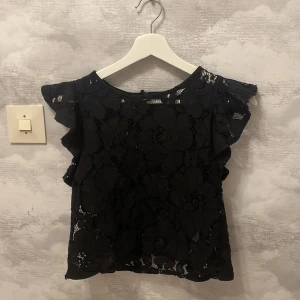Svart spetsblus från Zara Trafaluc - Säljer en elegant svart spetsblus från Zara Trafaluc. Blusen har volangdetaljer på ärmarna och ett vackert blommigt spetsmönster. Perfekt för vår och sommar.