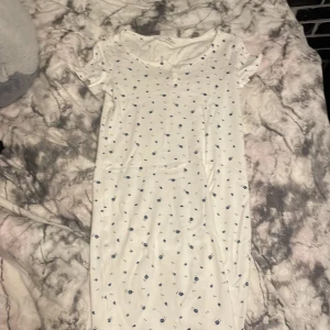 Vit kortklänning med blommigt mönster - Söt vit kortklänning med små blå blommor. Klänningen har korta ärmar och en rund halsringning. Perfekt för en avslappnad sommarlook.