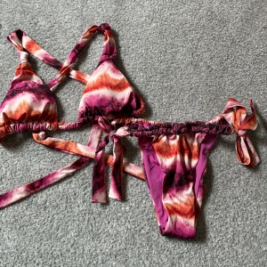 Färgglad bikini med knytband - Snygg bikini i härliga färger med mönster i rosa, orange och vitt. Toppen har knytband och korsade axelband för en unik look.  Man kan ändra om banden så den inte är korsade. Bikinitrosan har knytband i sidorna för justerbar passform. Perfekt för sommarens stranddagar! Super fin men har aldrig kommit till använding för mig. 