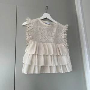 Topp med spets och volanger - Charmig krämvit topp från Zara med vackra spetsdetaljer och volanger. Toppen är ärmlös och har en luftig design med ett fint mönster på överdelen. Perfekt för en vår eller sommar look! Liten reva i spetsen som var där när jag köpte den tyvärr