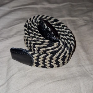 Svart och beige flätat bälte - Snyggt flätat bälte i svart och beige med ett zigzag-mönster. Bältet har svarta läderdetaljer vid ändarna och en klassisk spänne. Det är 110cm långt. Perfekt för att ge en stilren touch till din outfit. 