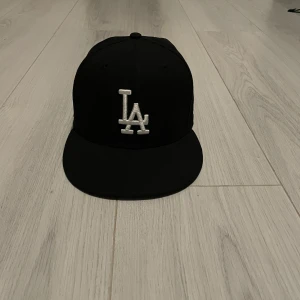 Svart keps med LA-logga - Snygg svart keps från New Era med vit LA-logga framtill och en guldkrona på sidan. Perfekt för att ge din outfit en sportig touch. Kepsen har en rak skärm och är i en klassisk stil.