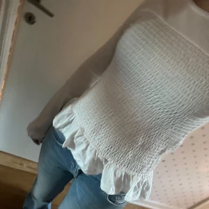 Vit smockad blus med volangdetaljer - Säljer en söt vit blus med smockad design och volangdetaljer i nederkant. Blusen har långa ärmar och en tight passform som ger en elegant look. Perfekt för både vardag och speciella tillfällen. Aldrig använd!!