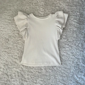 Vit ribbad topp från Zara - Säljer en stilren vit ribbad topp från Zara med volangdetaljer på ärmarna. Perfekt för en chic och avslappnad look. Toppen har en normal passform och är tillverkad i ett mjukt material.