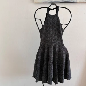 Svart glittrig klänning - Snygg svart glittrig klänning med en elegant halterneck-design. Klänningen har en kort längd och en utsvängd kjol som ger en fin siluett. Perfekt för en kväll ute eller en speciell tillställning. Den är stretchig. Från Pull & Bear. Mått: Längd fram: ca 79cm. Midjemått: 59cm men stretchig. Dragkedja: 17cm lång. 