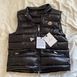 Svart dunväst från Moncler - Snygg svart dunväst från Moncler med dragkedja och två fickor framtill. Västen har en quiltad design och Monclers logotyp på bröstet. Perfekt för kyligare dagar när du vill hålla stilen.