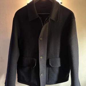 Mörkblå ulljacka med knappar från Massimo Dutti - Elegant mörkblå jacka i ull med knappar framtill och dragkedja och två stora fickor. Jackan har en klassisk krage och långa ärmar, perfekt för kyligare dagar.