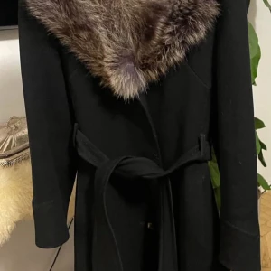 Svart ullkappa med päls från Distingue - Stilren svart kappa från Distingue med bred, lyxig päls runt kragen och knytskärp i midjan. Tillverkad i ren ny ull, perfekt för kalla dagar. Långärmad och elegant siluett som ger en classy vibe.