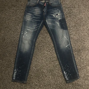 Dsquared2 Twinphony jeans med stänk - Snygga Dsquared2 Twinphony jeans i mörkblå tvätt med slitningar och vita färgstänk för en edgy look. Klassisk femficksmodell med smal passform och läderpatch bak i midjan. Perfekta för dig som gillar streetstyle och unika detaljer.