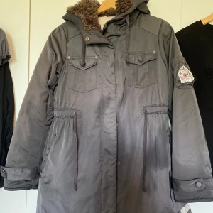 Grå parkasjacka från Odd Molly - Mysig grå parkasjacka från Odd Molly med brun teddyfoder i huva och längs insidan. Jackan har dragsko i midjan, stora fickor med lock och snygg patch på ena ärmen. Perfekt för kalla dagar med sin långa modell och varma känsla.