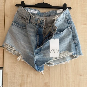 Högmidjade jeansshorts från Zara - Snygga ljusblå högmidjade jeansshorts från Zara med råa, fransiga benslut och slitna detaljer framtill. Klassisk femficksmodell med dragkedja och knapp. Perfekta för en chill sommardag och riktigt trendiga i modellen. 