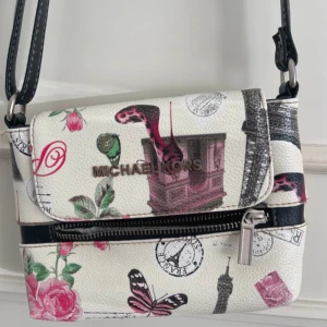 Michael Kors axelväska med Paris-motiv - Snygg axelväska från Michael Kors i vitt skinn med coolt Paris-inspirerat tryck. Väskan har detaljer som rosor, fjärilar, frimärken och Eiffeltornet. Justerbar svart axelrem och praktisk dragkedja framtill. Perfekt för dig som gillar unika accessoarer!