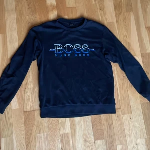 Mörkblå Hugo Boss sweatshirt slim fit - Snygg mörkblå sweatshirt från Hugo Boss med broderad logga i vitt och blått framtill. Tröjan har rund hals, långa ärmar och en slim fit passform. Perfekt för dig som gillar stilrena och klassiska plagg med en modern touch.