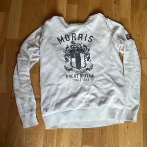 Vit stickad tröja från Morris S - Vit stickad tröja från Morris med tryck och emblem på ryggen samt Union Jack-detalj på ärmen. Klassisk rund halsringning och ribbade muddar. Perfekt för dig som gillar brittisk preppy stil och vill ha något stilrent men ändå med attityd.
