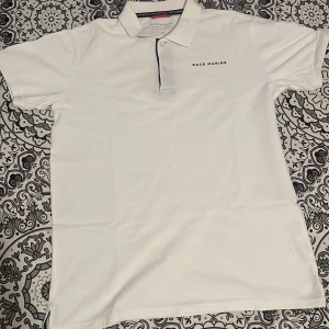 Vit pikétröja från Race Marine M - Säljer min klassiska vita pikétröja från Race Marine i storlek M. Tröjan har korta ärmar, krage och knäppning framtill med en diskret svart kant, perfekt för sommaren. Använd 2-3 gånger.