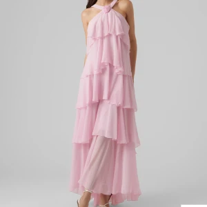 Ljusrosa volangklänning med öppen rygg - Superfin långklänning i ljusrosa med flera lager volanger och öppen rygg. Klänningen har breda axelband som korsas i nacken och en söt rosdetalj framtill. Tillverkad i ett lätt, flowy material som ger en drömmig känsla. Perfekt som sommarklänning eller som gäst på bröllop eller bal. Använd endast en gång, nypris 1200kr. Pris kan diskuteras.