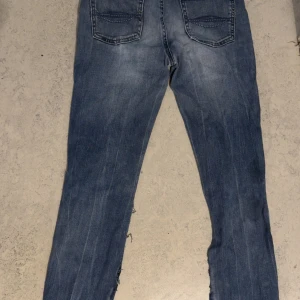 Blå jeans med råa slitsade ben - Säljer ett par blå jeans med slitningar och råa, öppna sidsömmar längs benen för en edgy look. Jeansen har klassisk femficksdesign, normal passform och är tillverkade i denim. Perfekta för dig som gillar en streetstyle vibe.