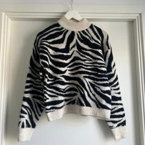 Zebramönstrad stickad tröja New Look - Säljer en stickad tröja från New Look i storlek S med svartvitt zebramönster. Tröjan har ribbad hög krage, muddar vid ärmslut och nederkant. Perfekt för dig som vill sticka ut med en cool och trendig look.