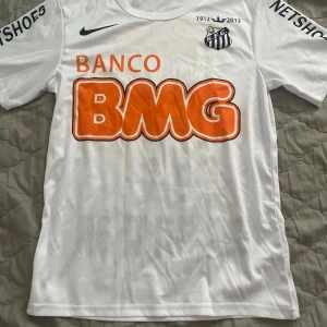Santos Neymar Jr fotbollströja Nike - Vit Santos fotbollströja från Nike med Neymar Jr och nummer 11 på ryggen. Tröjan har tryckta sponsorer i orange och svart, klubbmärke på bröstet och årtalet 1912-2012. Inte använd än enda gång. Priset kan diskuteras.