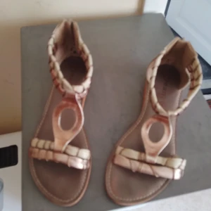 Beige och brons sandaler Cream Deluxe - Snygga sandaler från Cream Deluxe i beige och brons med flätade detaljer och öppen tå. Skorna har dragkedja bak för enkel påtagning och en platt sula. Perfekta för varma dagar när du vill ha en cool och unik look.