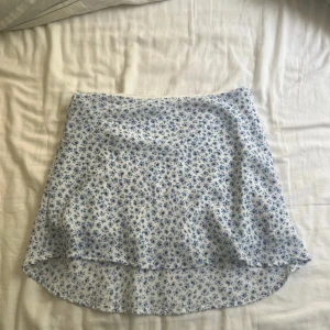 Blommig kortkjol från H&M Divided - Supersöt vit kortkjol med blått blommönster från H&M Divided. Kjolen har en lätt A-linjeform och är perfekt för varma dagar. Tunt och skönt material som känns luftigt och fräscht. Passar perfekt till en enkel topp och sneakers.