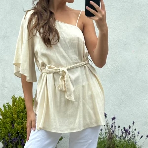 Beige oneshoulder linnetopp med knytband - Säljer en superfin beige linnetopp i oneshoulder-modell med en tunn axelrem och en lös, fladdrig ärm. Toppen har ett matchande knytband i midjan som ger en snygg siluett. Perfekt för varma dagar och riktigt skön mot huden.