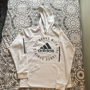 Vit hoodie från Adidas med tryck - Snygg vit hoodie från Adidas med stort svart tryck på bröstet och klassiska ränder på ärmarna. Tröjan har huva och en stor magficka framtill. Perfekt för en sportig och avslappnad stil. Skriv vid intresse/mer bilder!