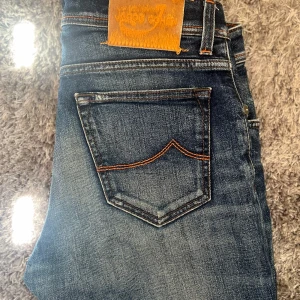 Blå jeans från Jacob Cohen - Snygga blå jeans från Jacob Cohen med klassisk femficksdesign och orange kontrastsömmar. Jeansen har en mellanblå tvätt med slitningar och en läderpatch bak i midjan. Tillverkade i mjukt denimtyg som sitter skönt hela dagen. Jeansen är omsydda så om du är 160cm passar de perfekt.