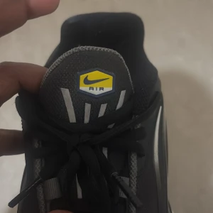 Nike Air Max Plus Tn svart/grå sneaker - Nike Air Max Plus Tn sneakers i svart och grått med vita detaljer och gula loggor. Snygg vågig design på sidorna, svarta skosnören och mesh- samt syntetmaterial. Ikonisk Tn Air-dämpning i sulan för extra komfort. Perfekt för dig som gillar streetwear.