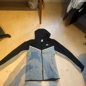 Nike svart och grå tech fleece S - Nike hoodiejacka i svart och grått med vit detalj över bröstet. Jackan har dragkedja, huva med snörning och två fickor framtill. Perfekt för chill eller träning, med klassisk Nike-logga på bröstet. Mjuk och bekväm i bomullsmix.