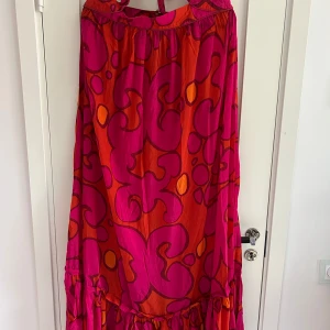 Marimekko x H&M långklänning mönstrad - Färgstark långklänning från Marimekko x H&M i storlek 42. Klänningen har ett unikt abstrakt mönster i rosa, rött och orange, med volang nertill och halterneck med knytning. Tillverkad i luftig bomull, perfekt för dig som vill sticka ut.