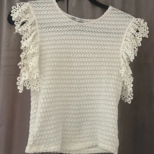 Vit spetsblus från Zara med volang - Supersöt vit blus från Zara med stickad struktur och detaljerade spetsvolanger på axlarna. Blusen är ärmlös och har en luftig känsla, perfekt för varmare dagar.💕☀️