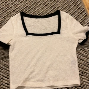Vit topp med svart kant och struktur - Vit kortärmad topp med fyrkantig ringning och svarta kanter vid hals och ärmslut. Tyget har en snygg strukturerad yta som ger extra detalj. Perfekt för dig som gillar stilrena kontraster och en unik twist på basplagget.