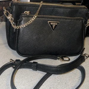 Svart Guess Noelle handväska - Säljer en svart Guess Noelle DBL Pouch Crossbody handväska med två fack, guldfärgade detaljer och kedja. Väskan har justerbar axelrem och Guess-logga framtill. Tillverkad i syntetmaterial med stilren struktur och dragkedja upptill.💕