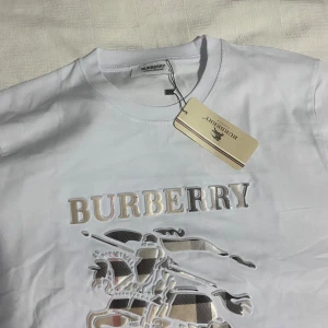 Vit Burberry t-shirt med logga - Vit t-shirt från Burberry med klassisk logga i metallic och broderad ryttare på bröstet. T-shirten har rund halsringning och korta ärmar. Materialet är mjuk bomull och trycket har inslag av Burberrys rutiga mönster.